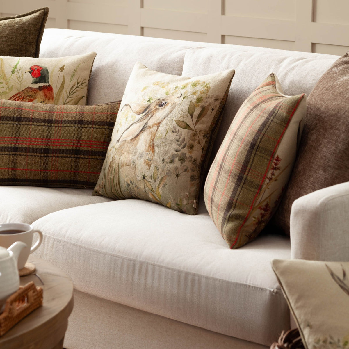 Bramble Hare Reversible Polyester Cushion in beige and green with a hand-painted hare and botanical print, styled on a cream sofa with plaid and pheasant-print cushions.