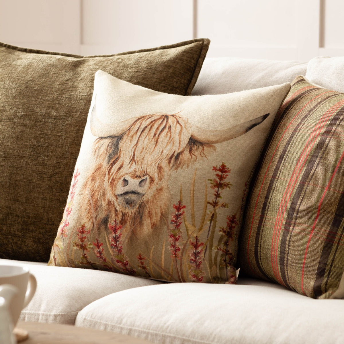 Bramble Highland Cow square cushion in beige polyester on a sofa, featuring a printed highland cow with shaggy amber fringe and rust-red wildflowers, styled with striped and olive scatter cushions.