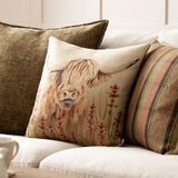 Bramble Highland Cow square cushion in beige polyester on a sofa, featuring a printed highland cow with shaggy amber fringe and rust-red wildflowers, styled with striped and olive scatter cushions.