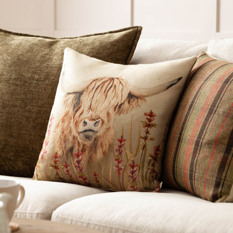 Bramble Highland Cow square cushion in beige polyester on a sofa, featuring a printed highland cow with shaggy amber fringe and rust-red wildflowers, styled with striped and olive scatter cushions.