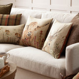 Bramble Highland Cow polyester cushion in beige with a printed highland cow and rust-red wildflowers, shown on a sofa among plaid and animal-print scatter cushions.
