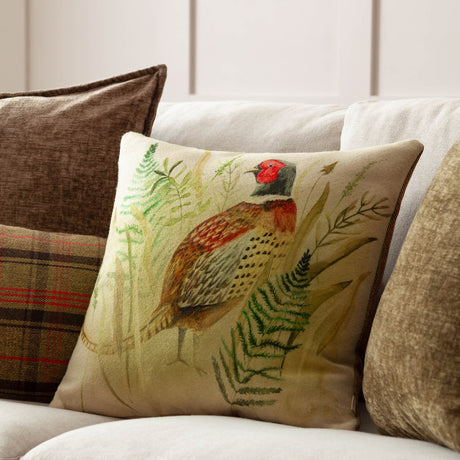 Bramble Pheasant square cushion in beige with a hand-painted pheasant among green ferns, shown on a sofa beside plaid and plain scatter cushions.