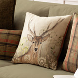 Square Bramble Stag cushion in cream and brown polyester with a hand-painted stag and meadow flowers, shown on an olive green sofa beside tartan cushions.
