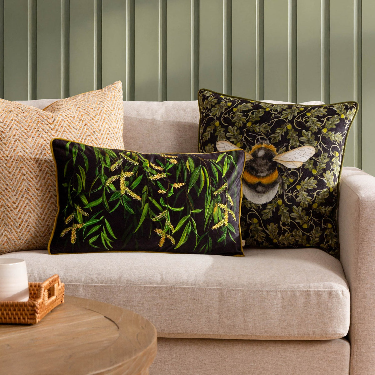 Braywick Animal Velvet Cushion in Bee design, a black and green square cushion with a printed bumblebee and oak leaf motif, shown on a beige sofa alongside patterned scatter cushions.
