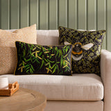 Braywick Animal Velvet Cushion in Bee design, a black and green square cushion with a printed bumblebee and oak leaf motif, shown on a beige sofa alongside patterned scatter cushions.