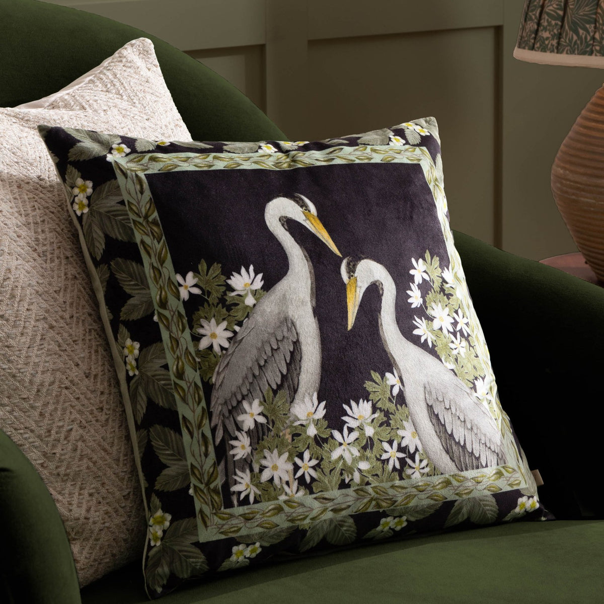 Braywick Animal Velvet Cushion in black velvet, printed with two grey cranes and amber beaks, framed by leaf and white floral border, styled on a green sofa.