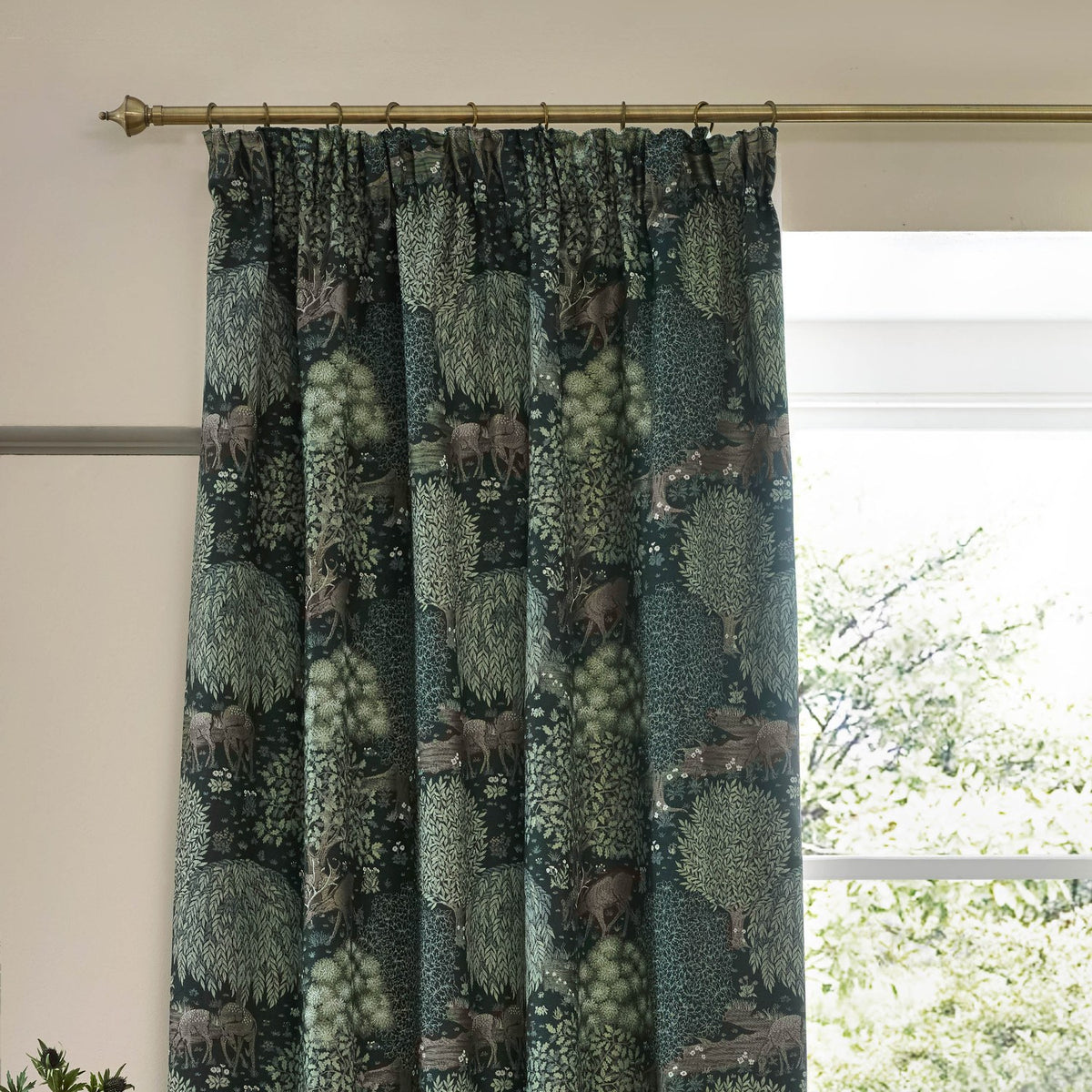 Brook Jacquard pencil pleat curtains in forest green polyester with woodland deer and tree pattern, hung on a brass rod beside a window.