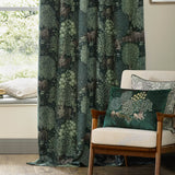 Brook Jacquard Polyester Pencil Pleat Curtains in Forest Green with woven woodland deer and tree motif, shown draped by a window beside a matching cushion on an armchair.