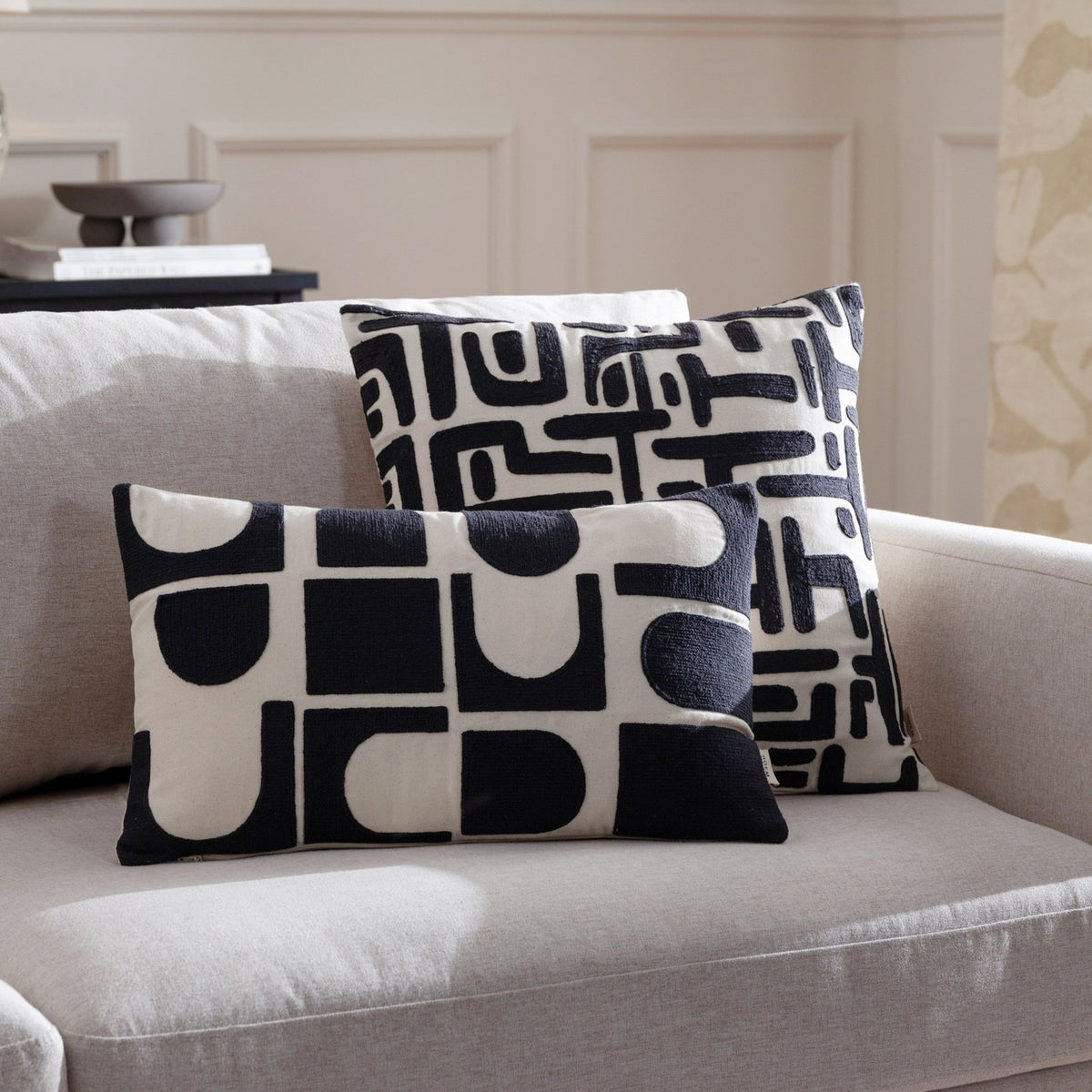 Brooke Abstract Cotton Cushion in Mono, a rectangular cream cotton cushion with black embroidered geometric shapes, shown with a matching square cushion on a light grey sofa.