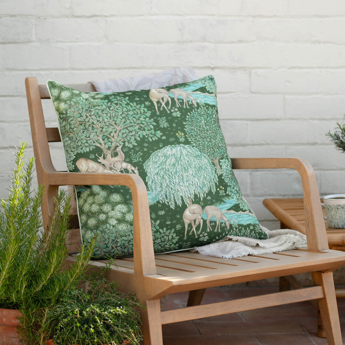 The Brook Jacquard Polyester Cushion in Forest Green on a wooden armchair, showing a woodland design with deer, oaks, and a willow tree by a brook.