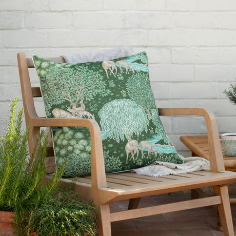 The Brook Jacquard Polyester Cushion in Forest Green on a wooden armchair, showing a woodland design with deer, oaks, and a willow tree by a brook.