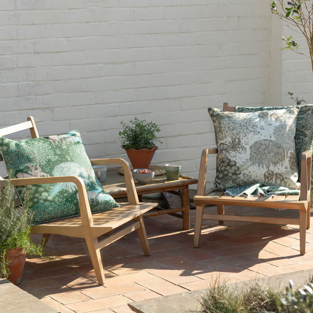 The Brook Jacquard Linen Cushion in green and cream, styled on wooden outdoor armchairs with a woodland deer print, shown on a sunlit patio.