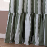 Birdie Stripe cotton pencil pleat curtains in bay green and mist, showing scalloped vertical stripes with a gathered ruffle skirt at the hem, against a pink wall.