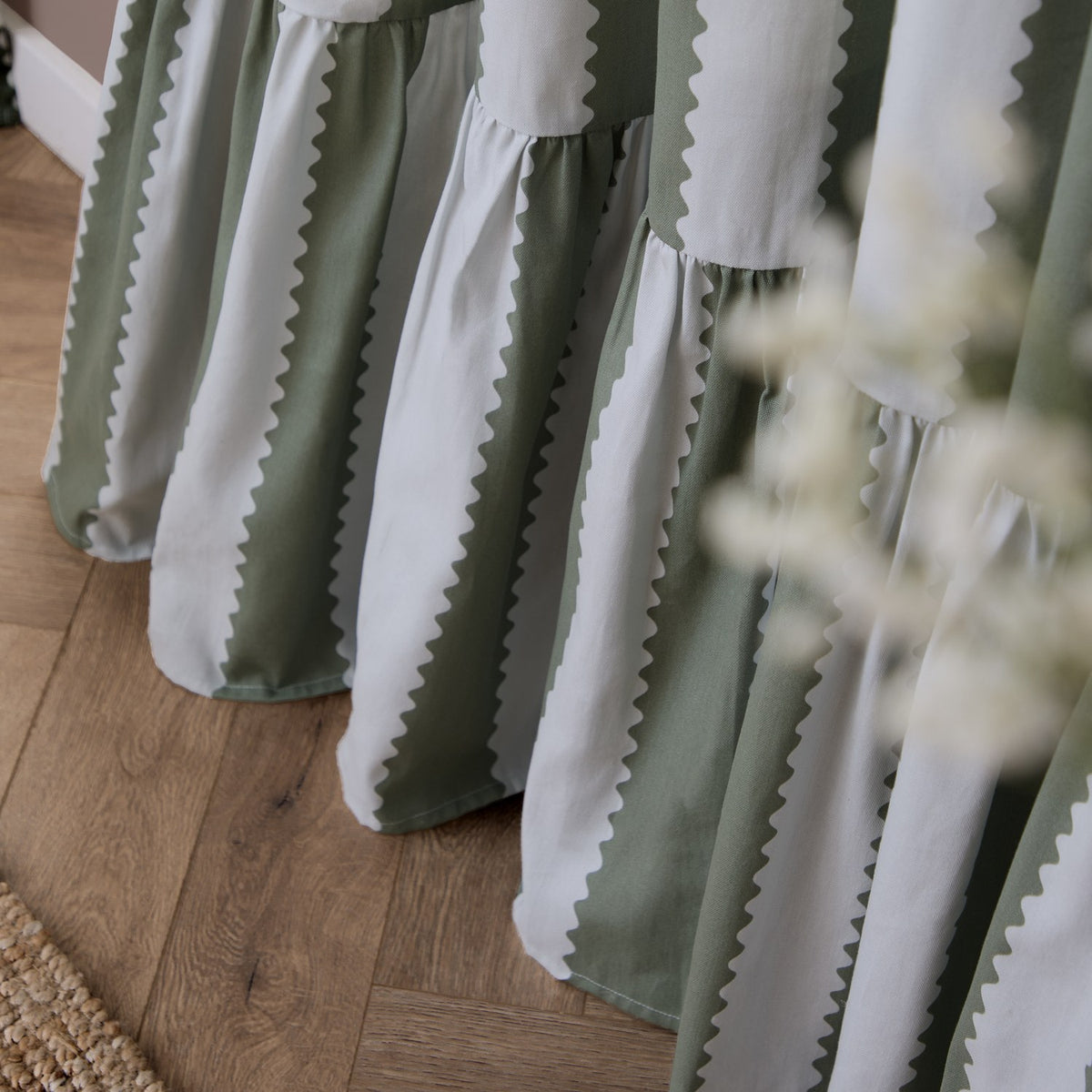 Close-up of Birdie Stripe pencil pleat curtains in bay green and mist, cotton fabric with wavy striped pattern and gathered ruffled hem, pooling on a wooden floor.