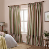 Birdie Stripe pencil pleat curtains in pink and green cotton stripes with a ruffled hem, hung on a wooden pole beside a shuttered window in a bedroom.