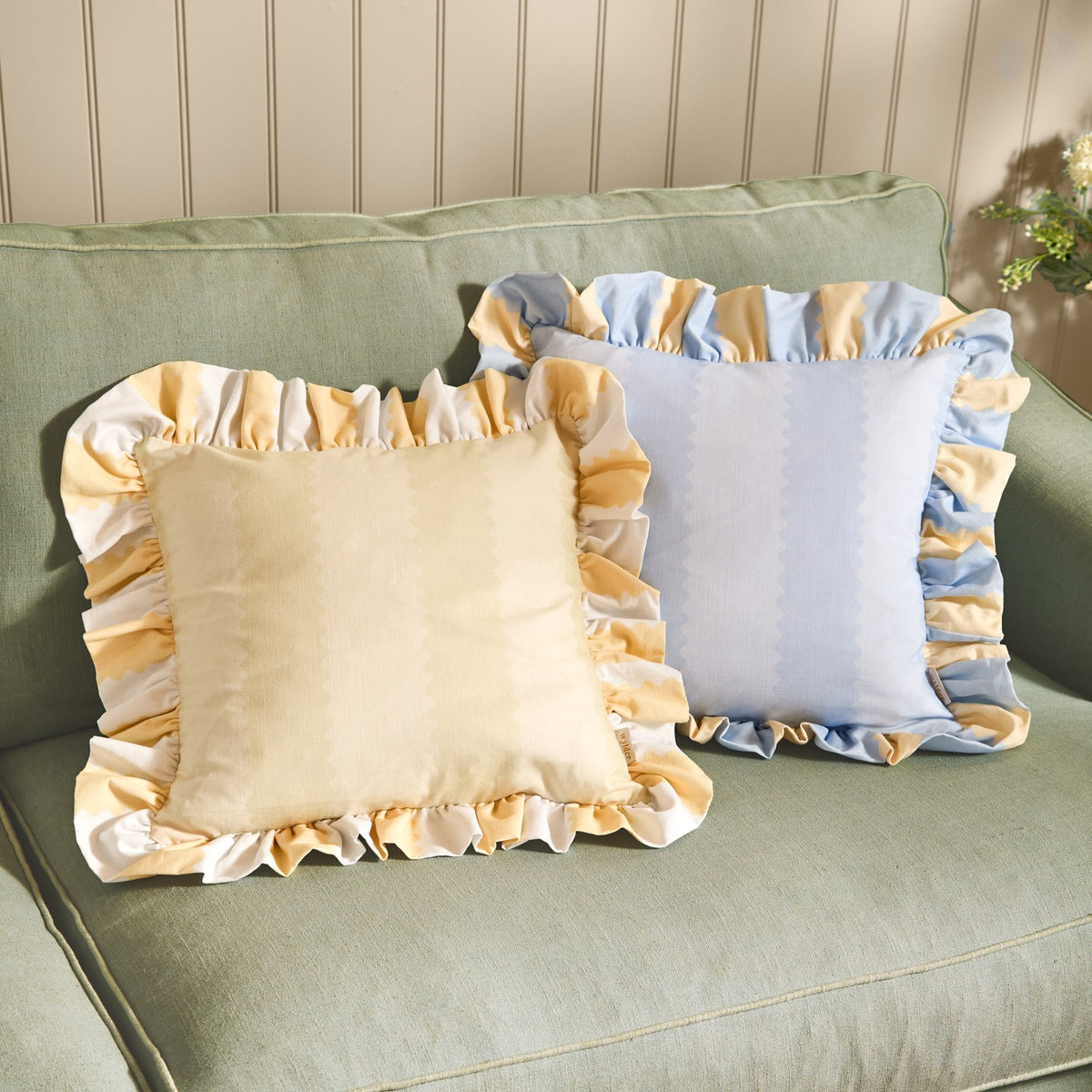 Two Birdie Stripe cotton cushions with gathered frill trim on a green sofa, one lemon with pale stripes, one sky blue with matching yellow-blue ruffle edging.