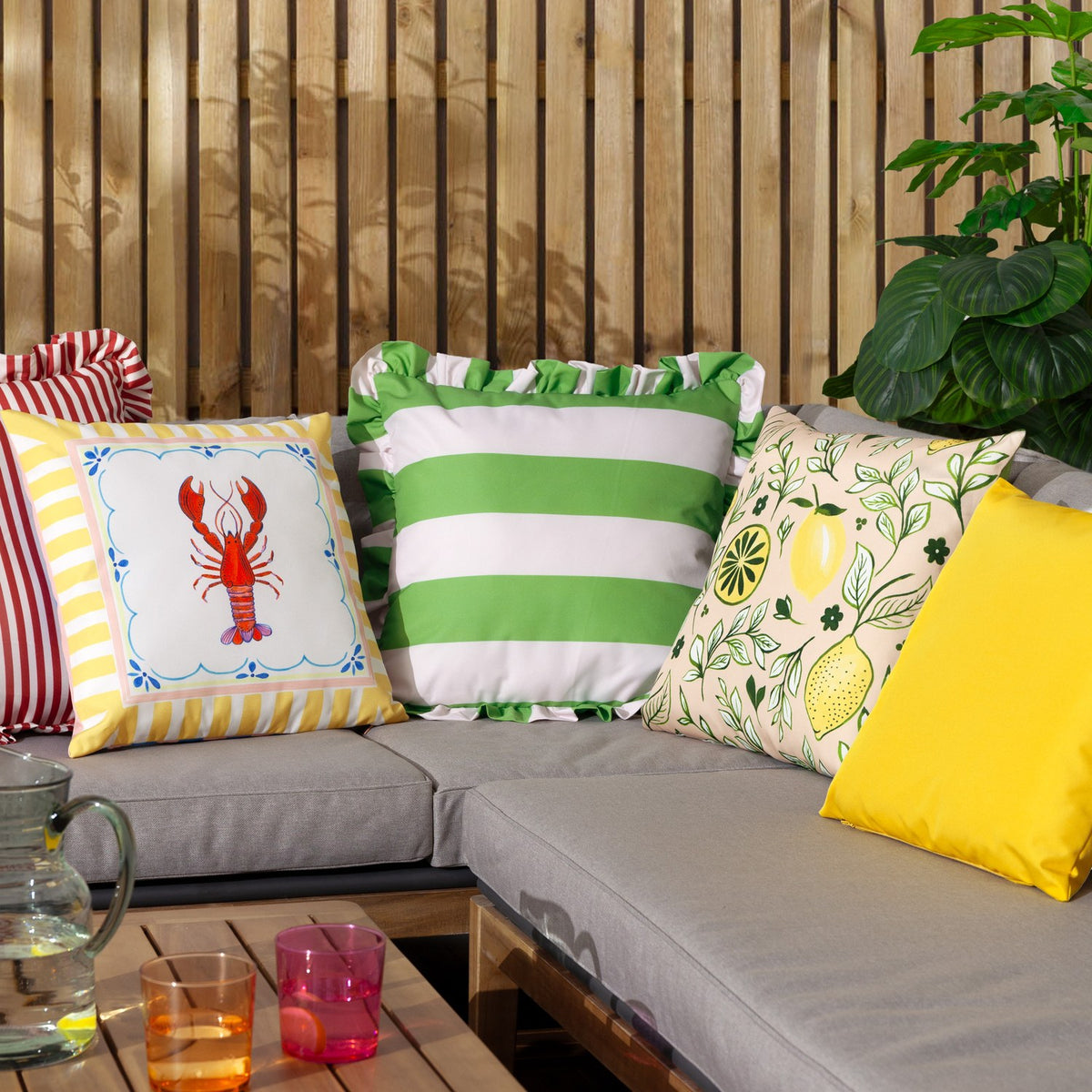 Brunch Stripes reversible polyester cushion in yellow-and-white stripe with a hand-painted lobster design, styled on an outdoor sofa among green striped and lemon-print scatter cushions.