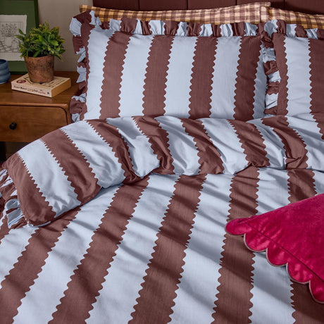 Blue and chocolate Birdie Stripe duvet cover set with wavy vertical stripes, zig-zag edging, and ruffled pillow sham trim, styled on a bed with a red scalloped cushion.