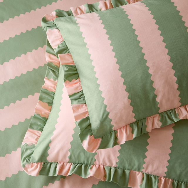 Birdie Stripe cotton blend pillowcase pair in pink and green, featuring a wavy scalloped stripe design with a gathered ruffle edge, shown layered on matching bedding.