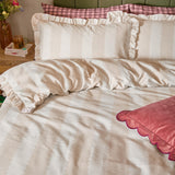 Birdie Stripe duvet cover set in oat and milk, styled on a bed with frill-trimmed pillowcases and a contrasting pink scalloped cushion.