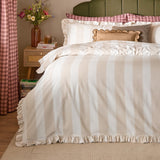 Birdie Stripe duvet cover set in oat and milk, with frill-trimmed edges and matching pillowcases, styled on a bed in a bedroom with checked textiles and a green headboard.