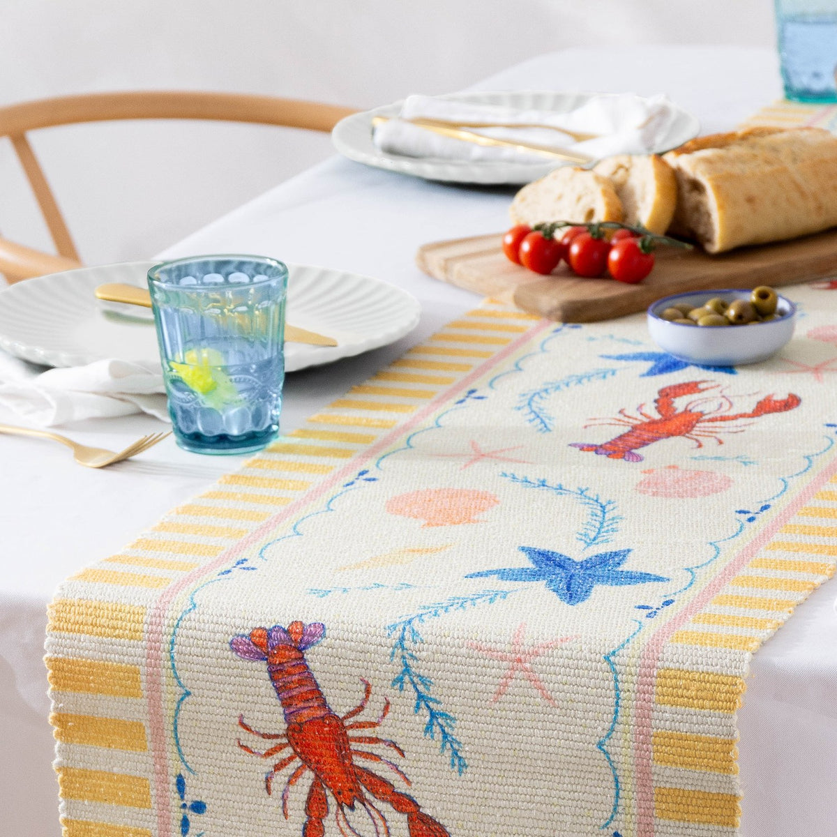 Brunch Stripes printed polyester table runner in cream with yellow striped borders, featuring lobsters, blue starfish and shells, styled on a dining table with bread, tomatoes and olives.