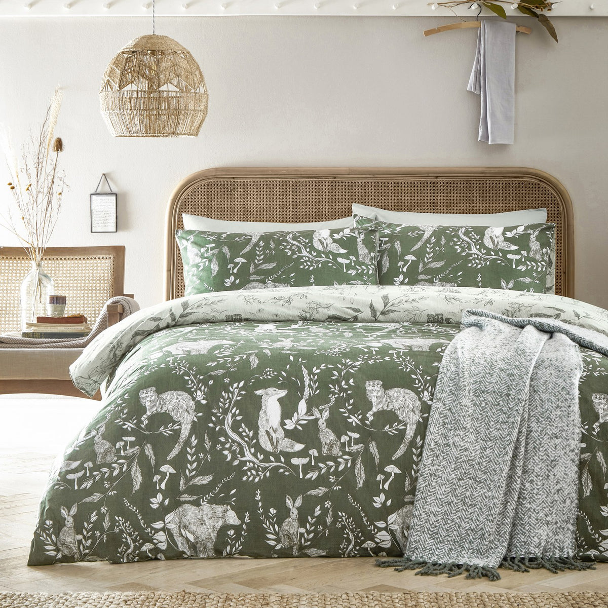 Buckthorn Reversible Animal Duvet Cover Set in Evergreen, styled on a bed with rattan headboard, showing woodland animals and foliage print with matching pillowcases and a grey throw.