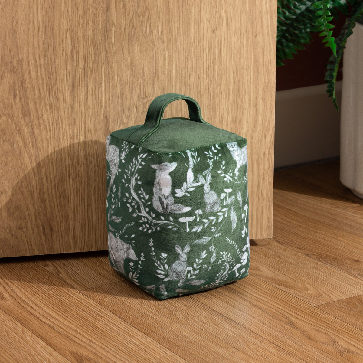 Buckthorn Animal Velvet Door Stop in evergreen, a fabric doorstop with woodland fox, rabbits and foliage print and a plain velvet top handle, shown by a wooden door on a hallway floor.