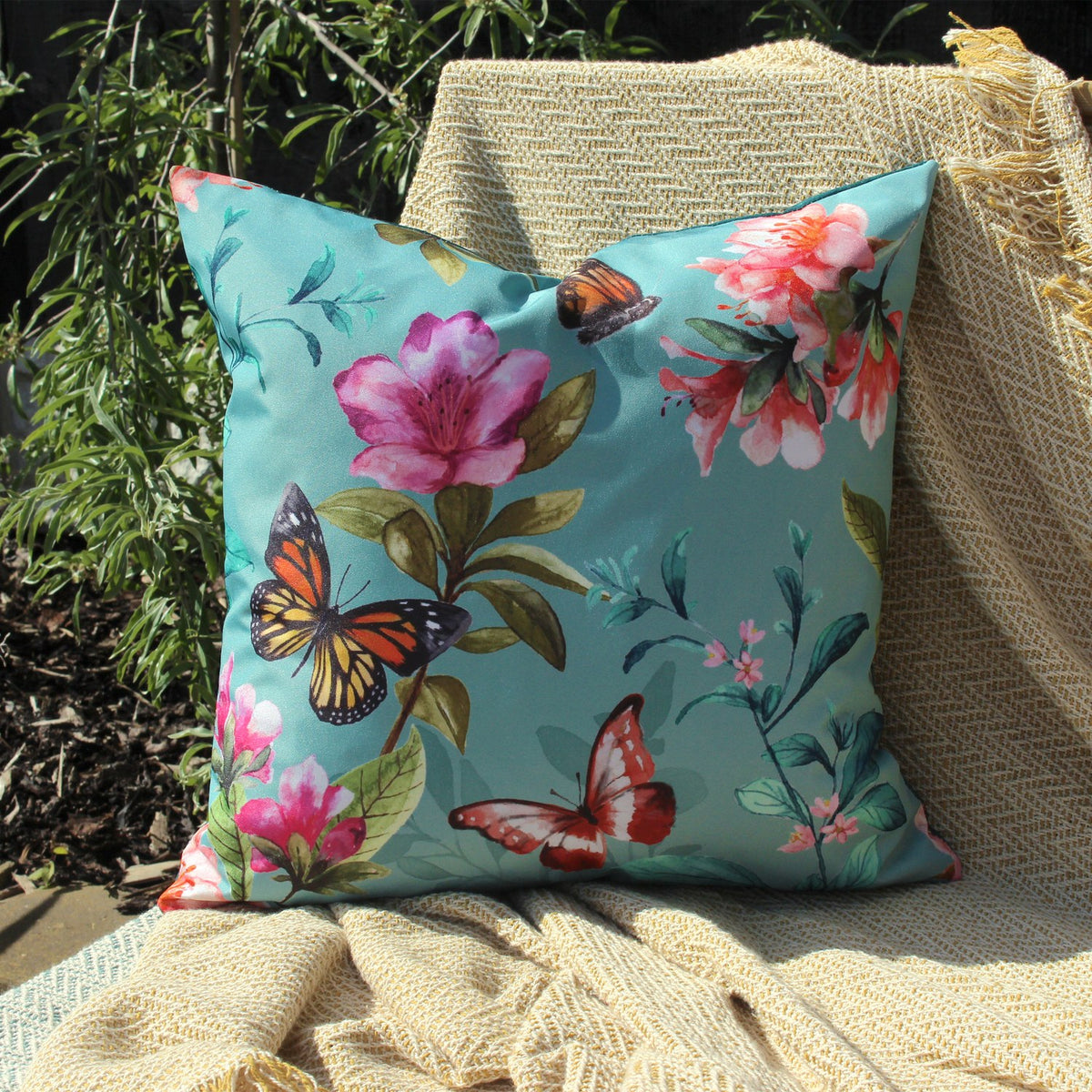 Square outdoor cushion in duck egg blue polyester with a watercolour print of pink and coral blooms, foliage and butterflies, shown outdoors on a woven chair with a cream throw.