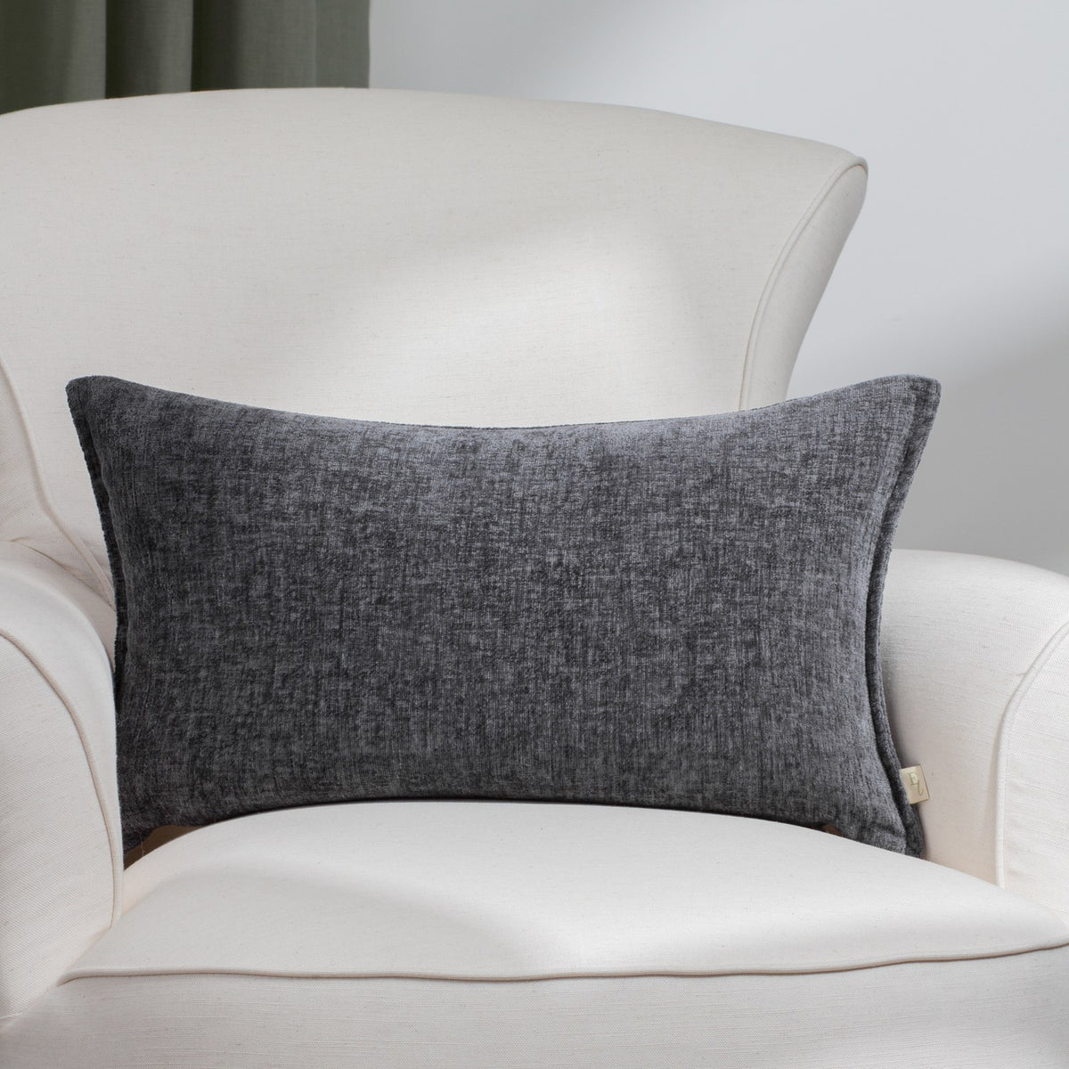 Buxton Reversible Polyester Cushion in charcoal grey, rectangular with a textured chenille-like weave, shown on a cream armchair in a living room setting.