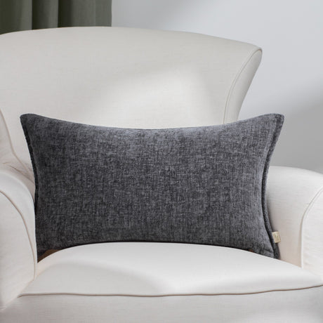 Buxton Reversible Polyester Cushion in charcoal grey, rectangular with a textured chenille-like weave, shown on a cream armchair in a living room setting.