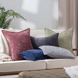 Buxton reversible chenille-texture polyester cushion in heather wine-rose, shown with navy, grey and green scatter cushions on a cream sofa in a living room setting.
