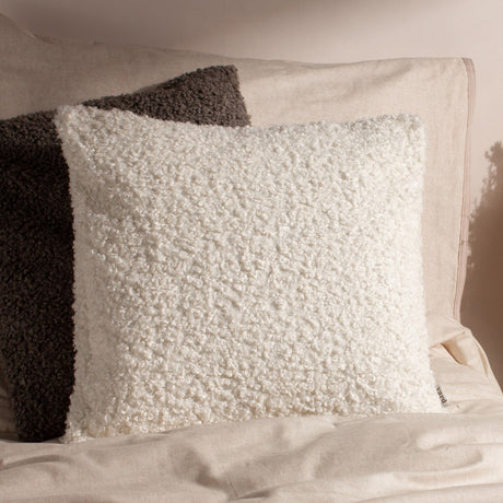 Cabu reversible wool cushion in cream with textured bouclé pile, shown square-shaped on a neutral bed setting beside a grey boucle pillow.