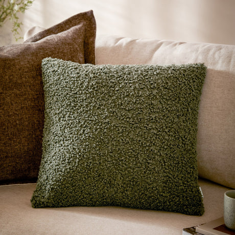 Cabu Reversible Wool Cushion in khaki, a square bouclé-textured cushion with a curled, fleece-like surface, shown on a sofa beside a brown cushion.