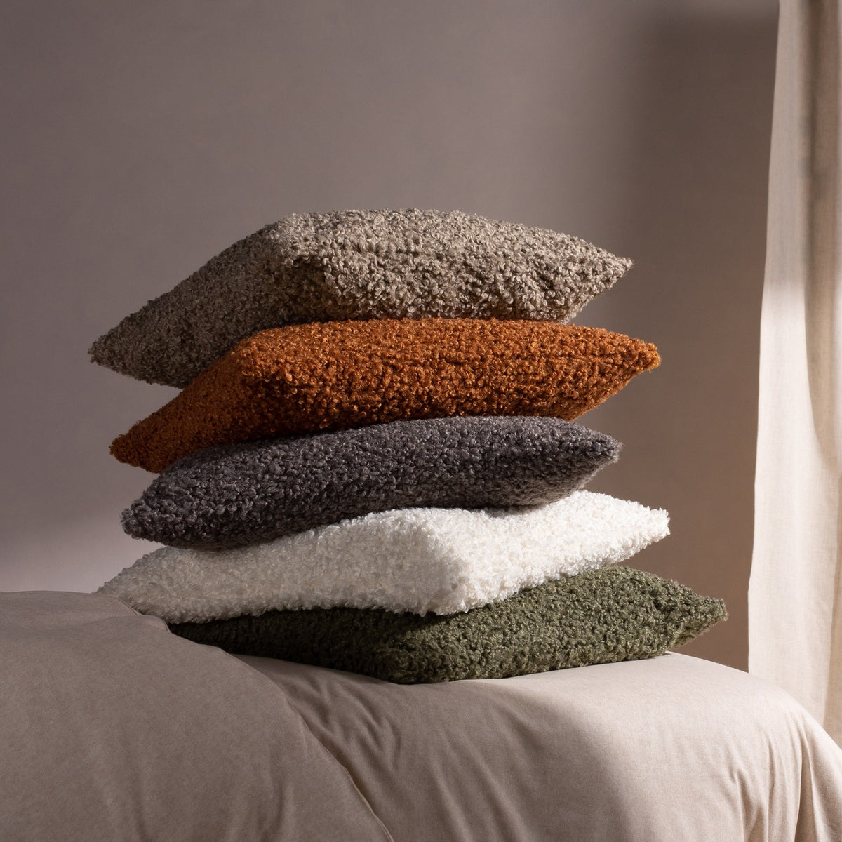 Cabu Reversible Wool Cushion in khaki, textured bouclé fabric, stacked with taupe, orange, grey and white cushions on a neutral bed setting.
