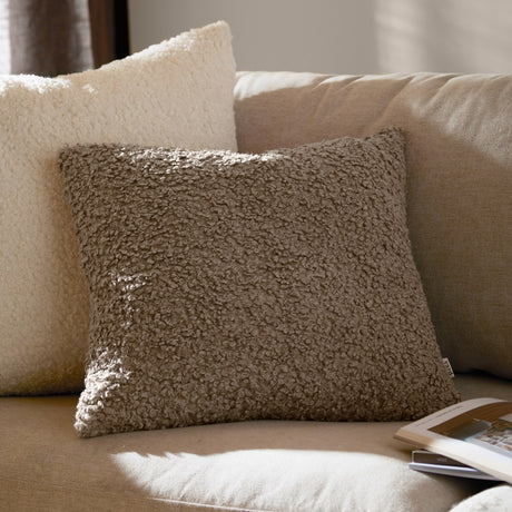 Cabu Reversible Wool Cushion in taupe, a square bouclé cushion with a curly shearling-look pile, styled on a beige sofa beside a cream boucle cushion.