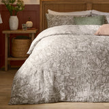 Cademuir Cotton Woodland Duvet Cover Set in nutmeg-brown with off-white toile print, shown on a bed with matching pillowcases, green and pink cushions, and a rattan headboard.