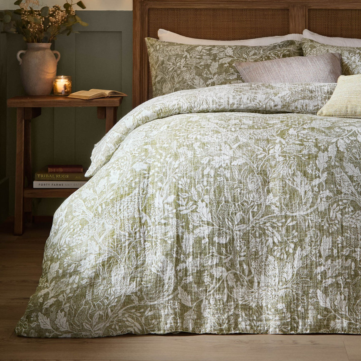 Cademuir Cotton Woodland Duvet Cover Set in sage, shown on a bed with matching pillowcases, featuring a white woodland toile print of birds, hares and leaves in a rustic bedroom setting.