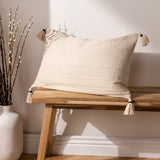 Caliche Plain Cotton Cushion in natural cream, showing textured woven stripes and corner tassels, styled on a wooden bench beside a vase of branches.