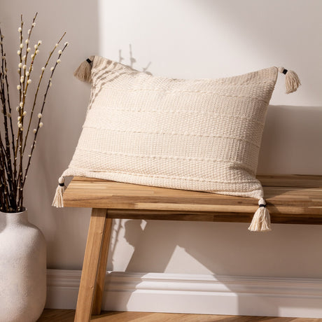 Caliche Plain Cotton Cushion in natural cream, showing textured woven stripes and corner tassels, styled on a wooden bench beside a vase of branches.