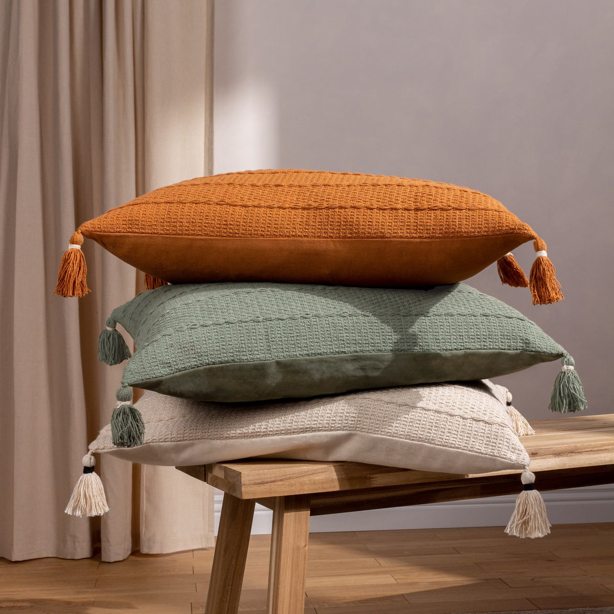 Caliche Plain Cotton Cushion in natural cream with tasselled corners, shown atop stacked green and orange textured cushions on a wooden bench.