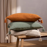 Caliche Plain Cotton Cushion in natural cream with tasselled corners, shown atop stacked green and orange textured cushions on a wooden bench.
