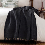 Candar Plain Polycotton Throw in black, boucle-textured with hand-knotted fringe, draped over a white bed in a bedroom setting.