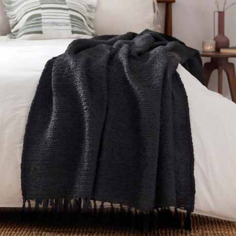 Candar Plain Polycotton Throw in black, boucle-textured with hand-knotted fringe, draped over a white bed in a bedroom setting.