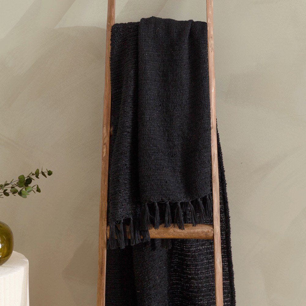 Candar Plain Polycotton Throw in black draped over a wooden ladder, showing its boucle-woven texture and hand-knotted fringe edge.