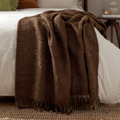 Candar Plain Polycotton Throw in cocoa brown, a boucle-woven textured blanket with knotted fringe ends, draped over a white bed in a bedroom setting.