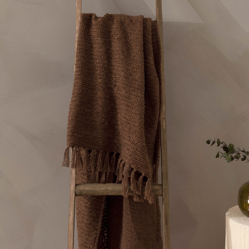 Candar Plain Polycotton Throw in cocoa brown, a boucle-woven blanket with knotted fringe ends, draped over a wooden ladder against a neutral wall.