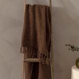 Candar Plain Polycotton Throw in cocoa brown, a boucle-woven blanket with knotted fringe ends, draped over a wooden ladder against a neutral wall.