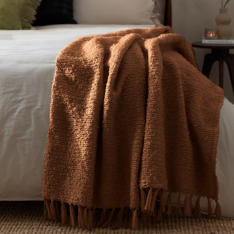 Candar Plain Polycotton Throw in ginger, a boucle-textured brown throw with fringed ends, draped over a white bed beside a wooden side table.