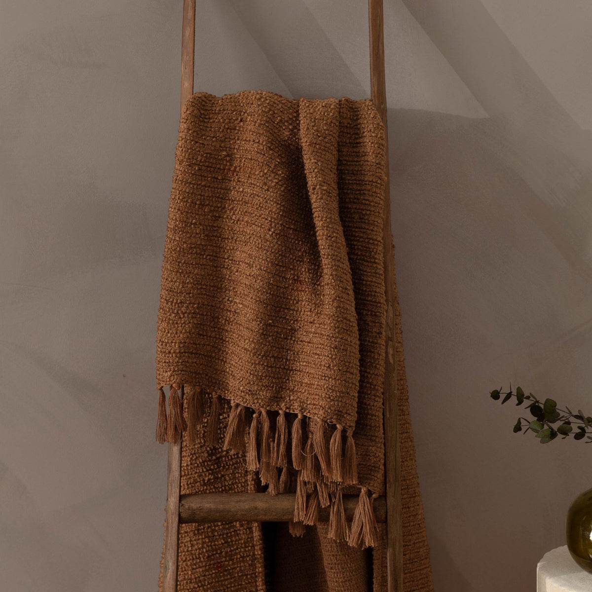 Candar Plain Polycotton Throw in ginger, a boucle-textured brown throw with hand-tied fringe, draped over a wooden ladder in a living room setting.
